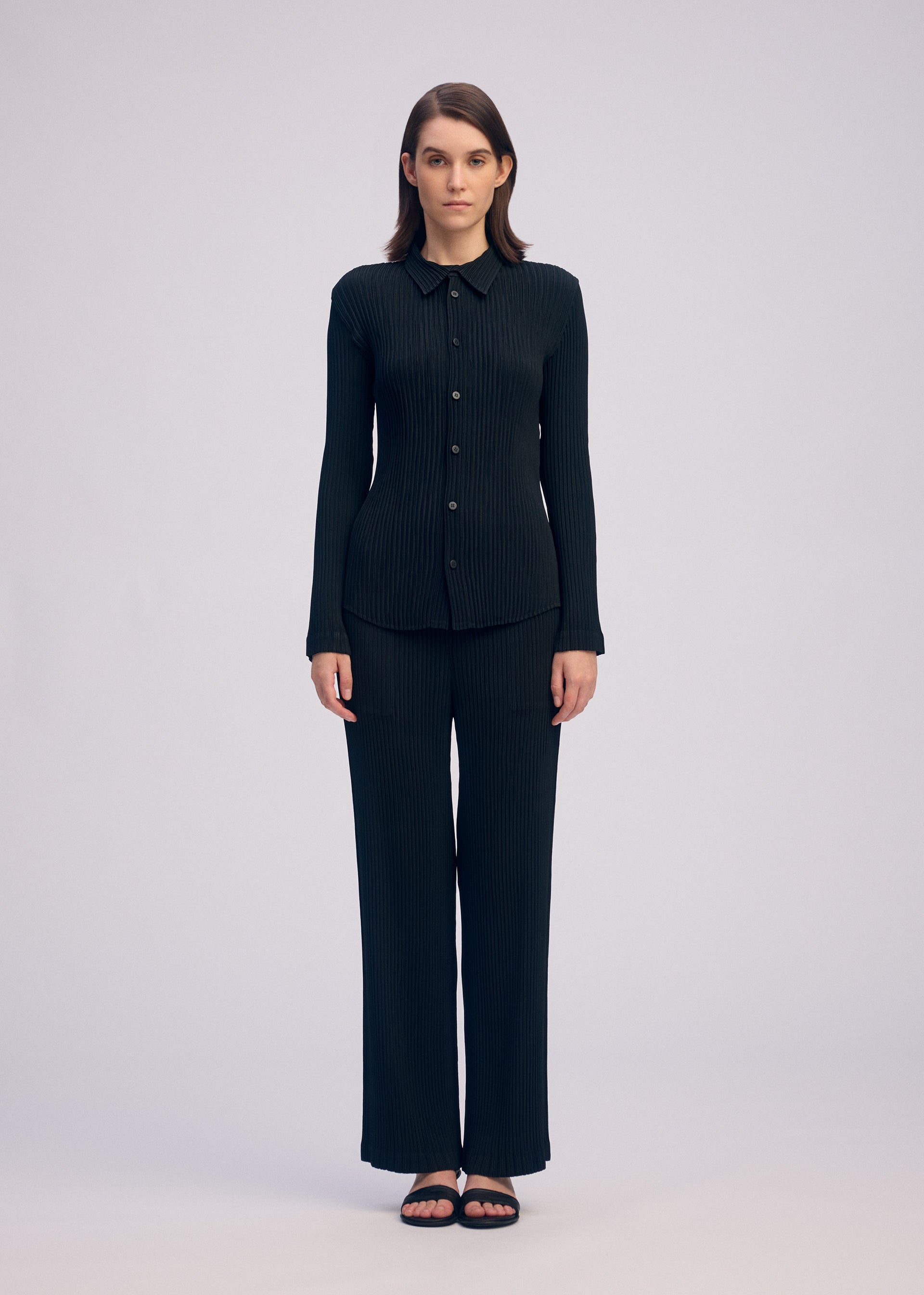 Zoomed view A woman with straight brown hair faces forward against a plain background, wearing the ISSEY MIYAKE HATCHING PLEATS black long-sleeve button-up top and matching pleated straight pants in recycled polyester, paired with black sandals.
