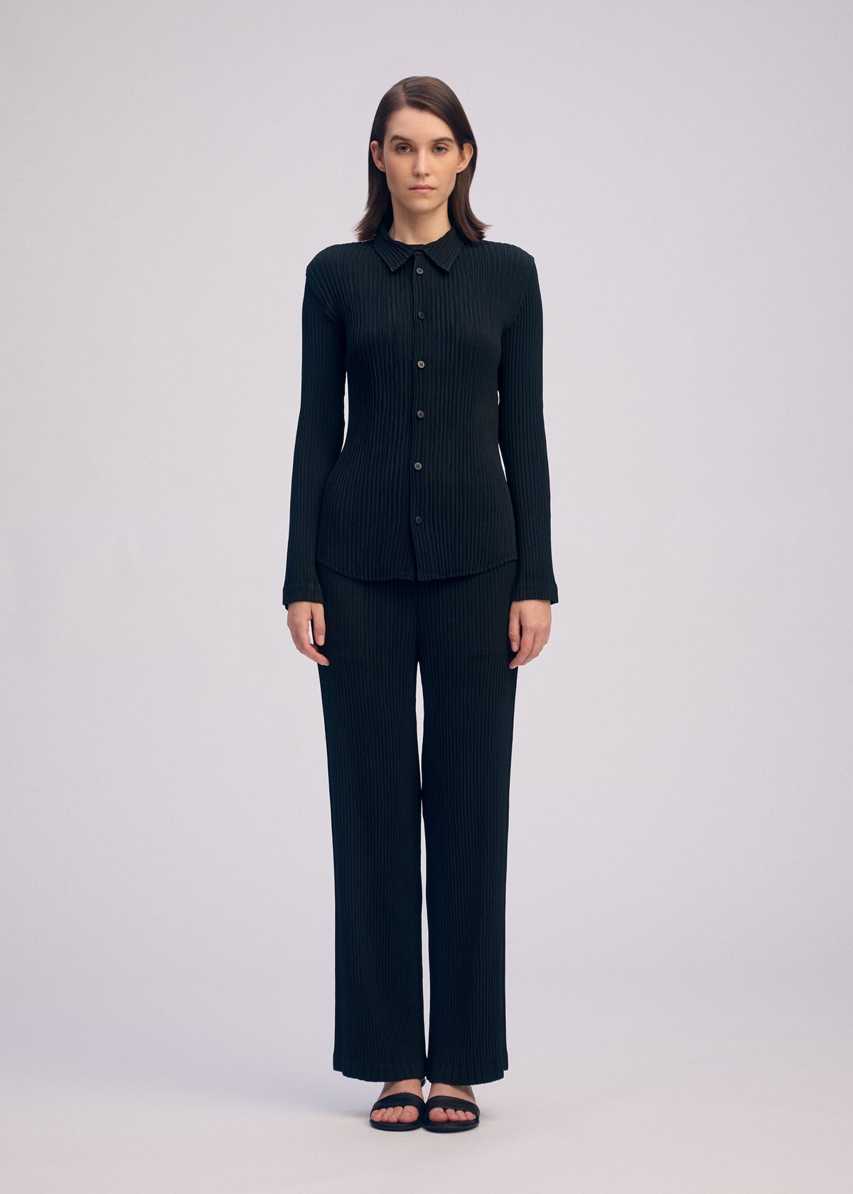 A woman faces forward against a plain background, wearing the ISSEY MIYAKE HATCHING PLEATS black long-sleeved button-up shirt and matching pleated straight pants in recycled polyester, paired with black sandals. Her arms are at her sides and she has a neutral expression. view 4 of 5