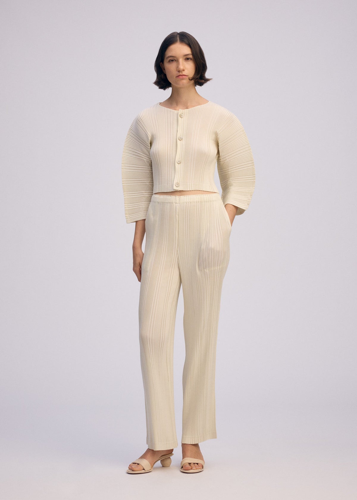 A woman with short dark hair wears ISSEY MIYAKE’s LINEN LIKE RIB PLEATS set—cream, textured, buttoned top with matching straight pants—against a plain background. She pairs it with open-toed sandals and has a neutral expression. view 7 of 8