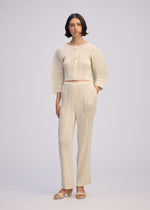 A woman with short dark hair wears ISSEY MIYAKE’s LINEN LIKE RIB PLEATS set—cream, textured, buttoned top with matching straight pants—against a plain background. She pairs it with open-toed sandals and has a neutral expression.