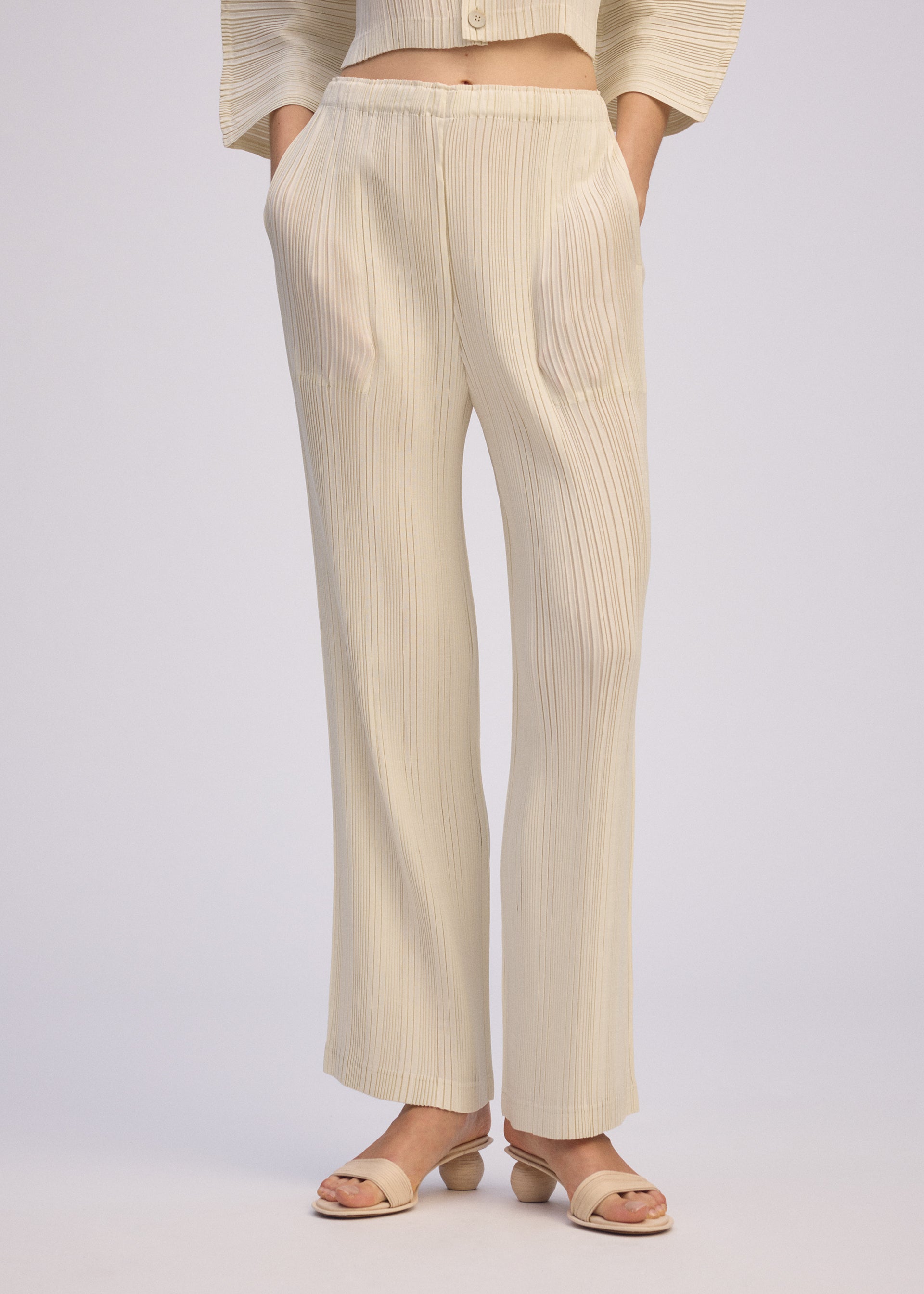 Zoomed view A person wears ISSEY MIYAKE's LINEN LIKE RIB PLEATS in light beige—loose, straight pleated pants with hands in pockets and matching open-toe sandals. Made from linen-like polyester, the cropped image captures a relaxed, casual look.