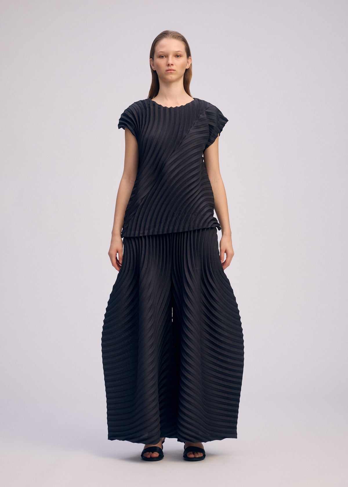 A person faces forward in the CIRCLE PLEATS PB set by ISSEY MIYAKE, featuring a matching black pleated top and wide pants made from plant-derived polyester with handmade pleats, styled minimally against a plain light gray background. view 6 of 7