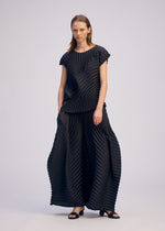 A woman in ISSEY MIYAKE's CIRCLE PLEATS PB stands before a plain background, wearing wide pants and a loose dark top with handmade pleats from plant-derived polyester, paired with straight hair and black sandals for a modern, simple look.