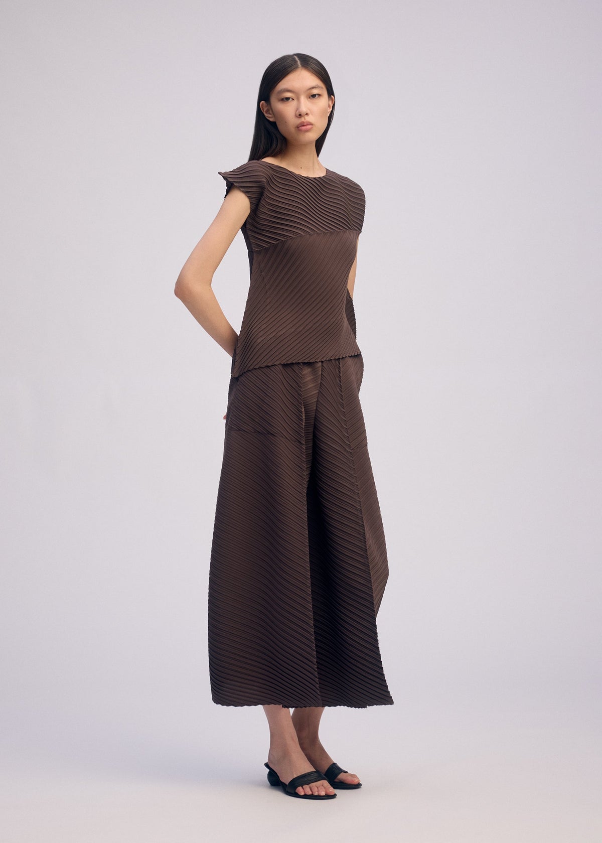 A woman with straight dark hair and black sandals stands against a plain background, wearing ISSEY MIYAKE's PLASTER PLEATS SOLID: wide pants and a matching dark brown recycled polyester crêpe de Chine top with subtle diagonal stripes. view 7 of 8