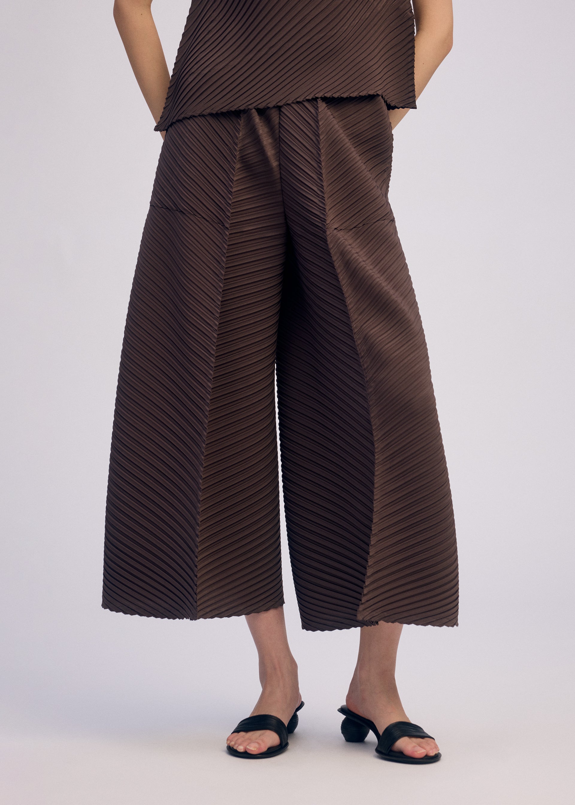 Zoomed view A person wears ISSEY MIYAKE PLASTER PLEATS SOLID wide-legged brown pleated pants and a matching diagonally striped top, styled with black open-toed sandals. The photo, shot against a plain background, is cropped from the shoulders down.