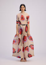 A woman wears ISSEY MIYAKE’s VERVE dress, featuring a beige base with red, blue, and black hand-drawn florals, short sleeves, fitted top, flared pleated skirt, and a black waist belt against a plain background.