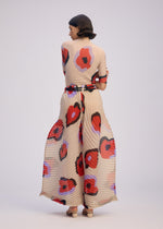 A woman faces away from the camera wearing the ISSEY MIYAKE VERVE set—beige pleated wide pants and a matching hand-drawn floral top, finished with a coordinating belt and cream heels—all against a light backdrop.