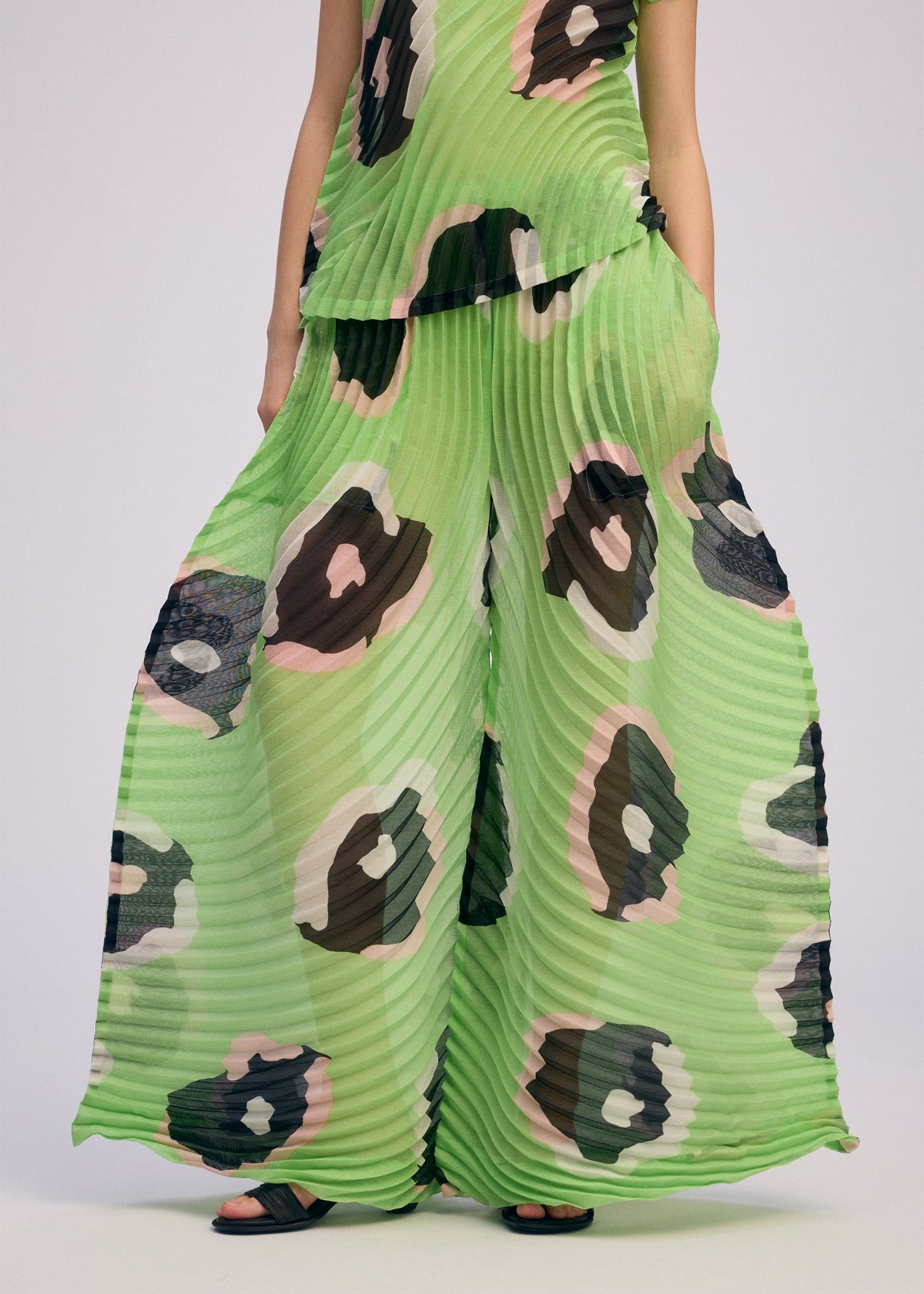 A person wears ISSEY MIYAKE's VERVE pleated wide pants, featuring a bold green, black, and pink abstract spot print. The handmade pleats stand out against a plain background, highlighting the unique design. view 9 of 10