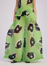 A person wears ISSEY MIYAKE's VERVE pleated wide pants, featuring a bold green, black, and pink abstract spot print. The handmade pleats stand out against a plain background, highlighting the unique design.