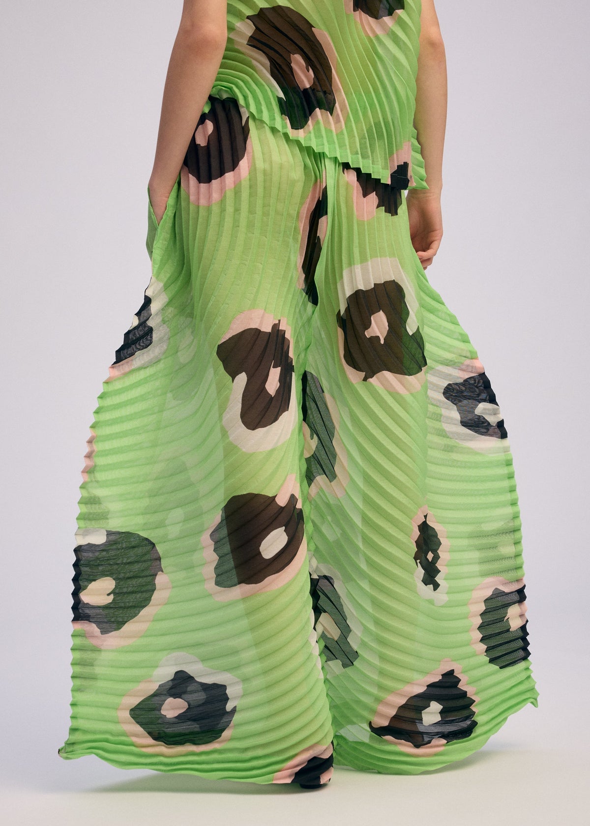 A person wearing ISSEY MIYAKE's VERVE pleated wide pants in semi-sheer lime green with hand-drawn brown, black, and pale pink florals stands hands-in-pockets, photographed from mid-torso down. view 10 of 10