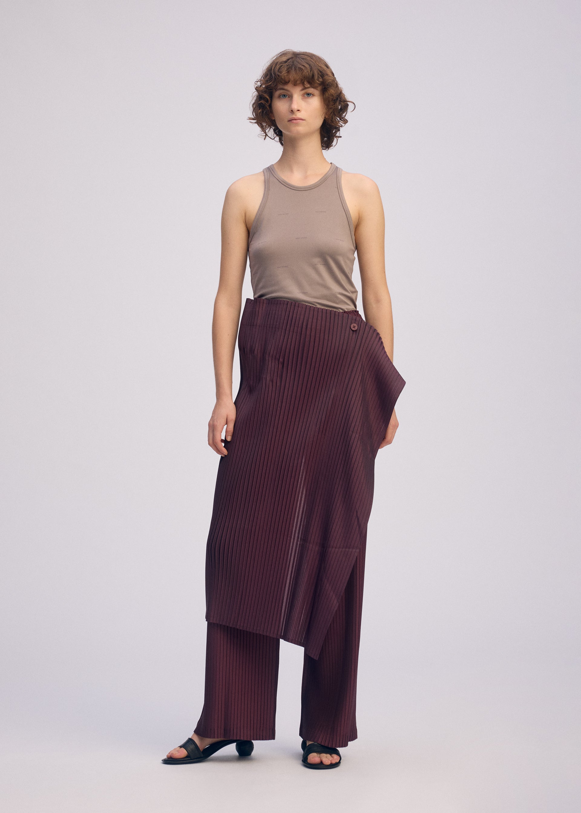 Zoomed view A person with short curly hair models the ISSEY MIYAKE PLEATED ON PLEATS wide-legged maroon pinstripe pants, paired with a sleeveless beige top and black sandals against a plain light background.