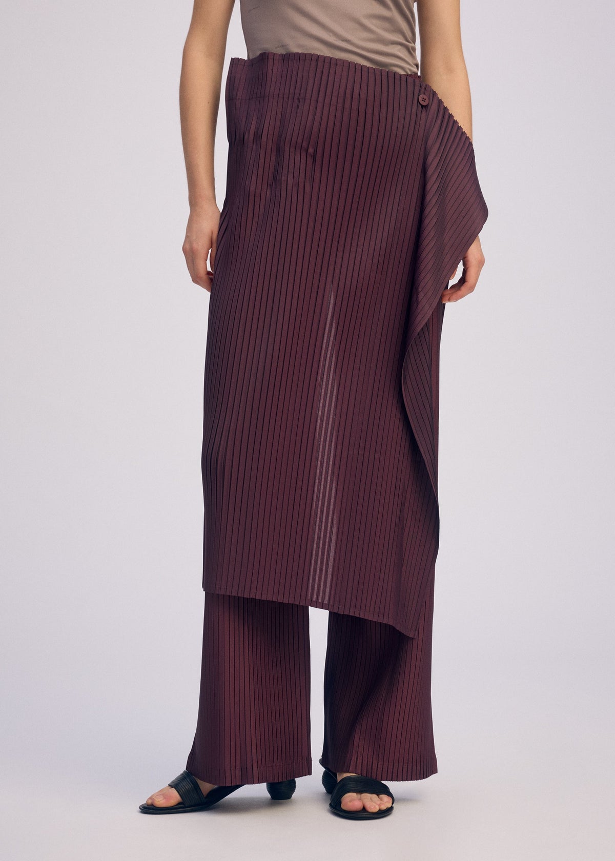 A person wears the ISSEY MIYAKE PLEATED ON PLEATS set—taupe sleeveless top with burgundy pinstriped layered and pleated trousers, plus black sandals—standing against a plain light background. view 8 of 8