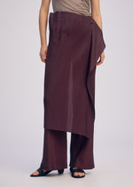 A person wears the ISSEY MIYAKE PLEATED ON PLEATS set—taupe sleeveless top with burgundy pinstriped layered and pleated trousers, plus black sandals—standing against a plain light background.