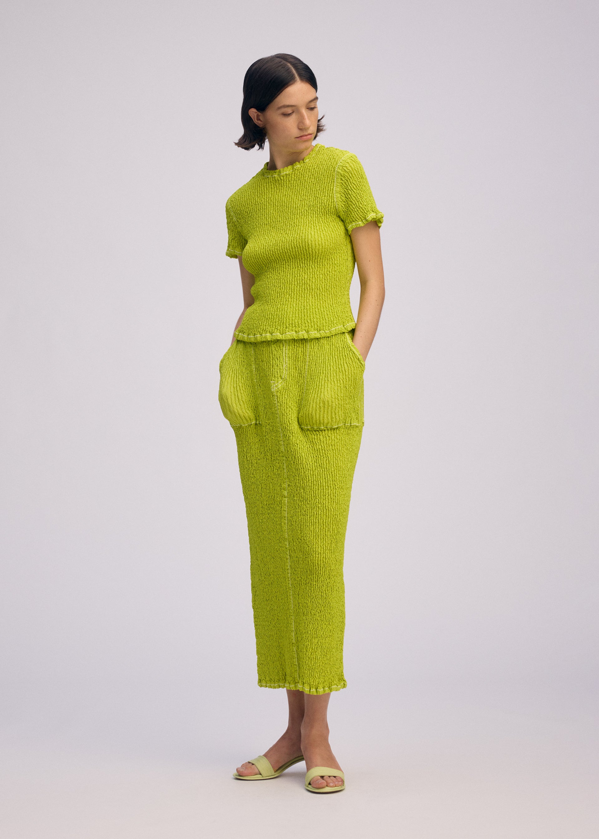 Zoomed view A woman with short dark hair stands against a plain light background, wearing the WAVELET ENSEMBLE by ISSEY MIYAKE: a bright lime green jacquard stretch knit top and matching long skirt with front pockets, paired with green sandals.