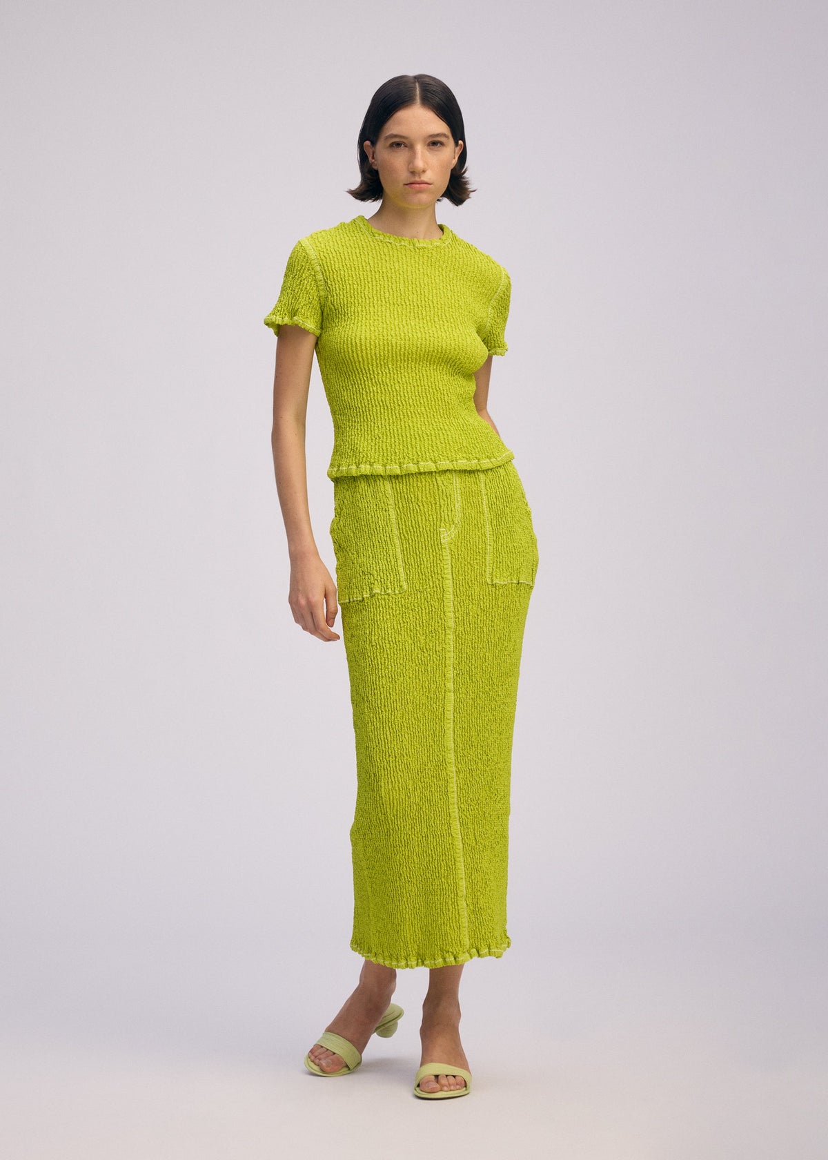 A woman with short dark hair and a neutral expression models the ISSEY MIYAKE WAVELET ENSEMBLE— a bright yellow-green jacquard stretch knit top and matching long skirt with pockets—against a plain background, paired with open-toe sandals. view 8 of 9