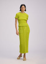A woman with short dark hair and a neutral expression models the ISSEY MIYAKE WAVELET ENSEMBLE— a bright yellow-green jacquard stretch knit top and matching long skirt with pockets—against a plain background, paired with open-toe sandals.