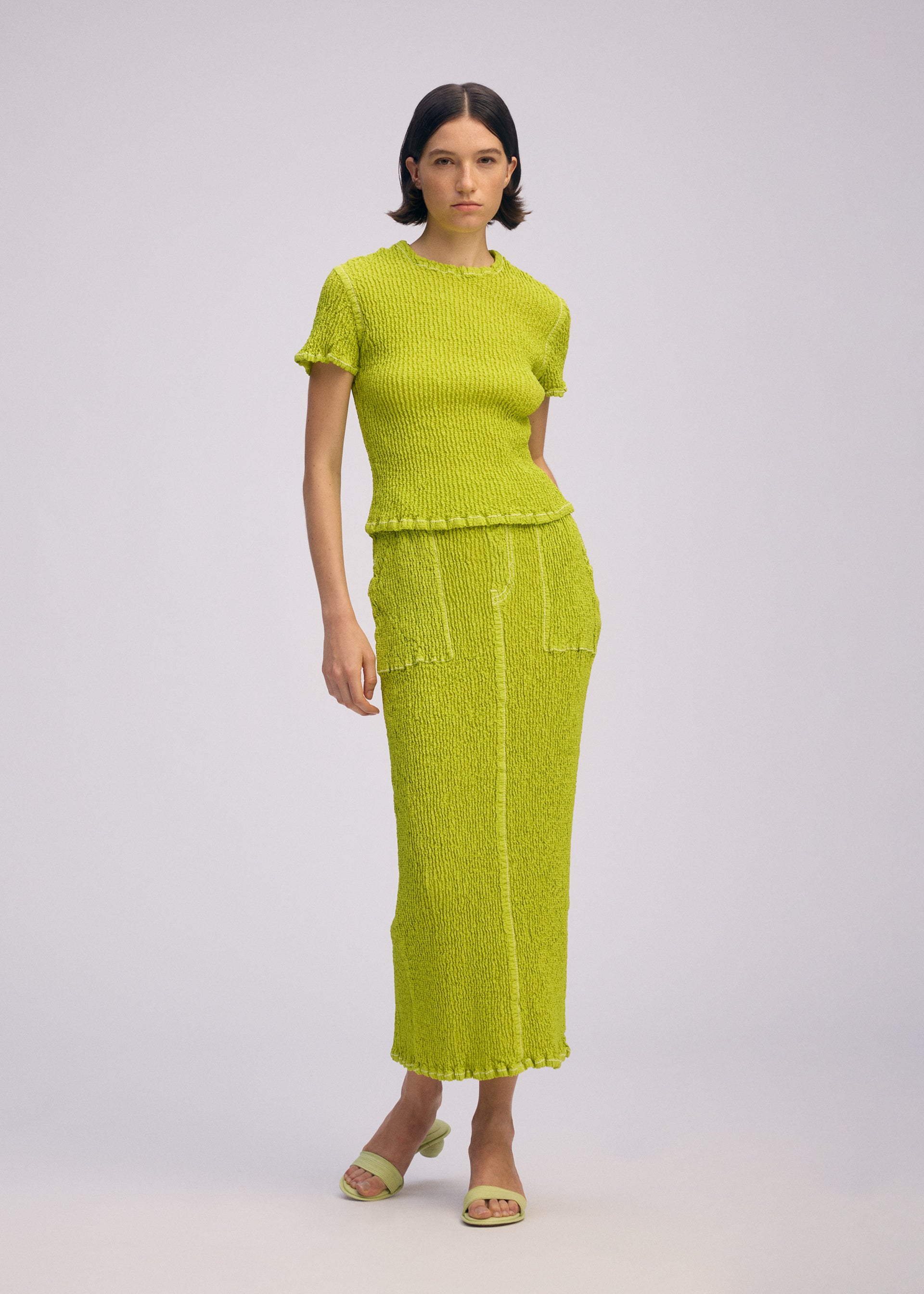 Zoomed view A woman with short dark hair and a neutral expression models the ISSEY MIYAKE WAVELET ENSEMBLE— a bright yellow-green jacquard stretch knit top and matching long skirt with pockets—against a plain background, paired with open-toe sandals.
