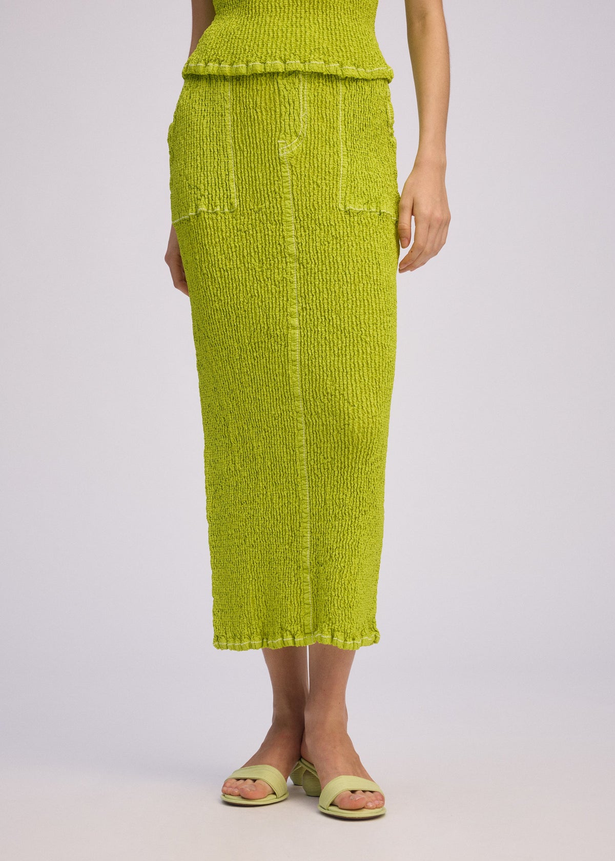 A person wears the ISSEY MIYAKE WAVELET ENSEMBLE—a textured, bright lime green jacquard stretch midi skirt and matching sleeveless top with open-toe sandals; only the lower half is visible against a light background. view 9 of 9