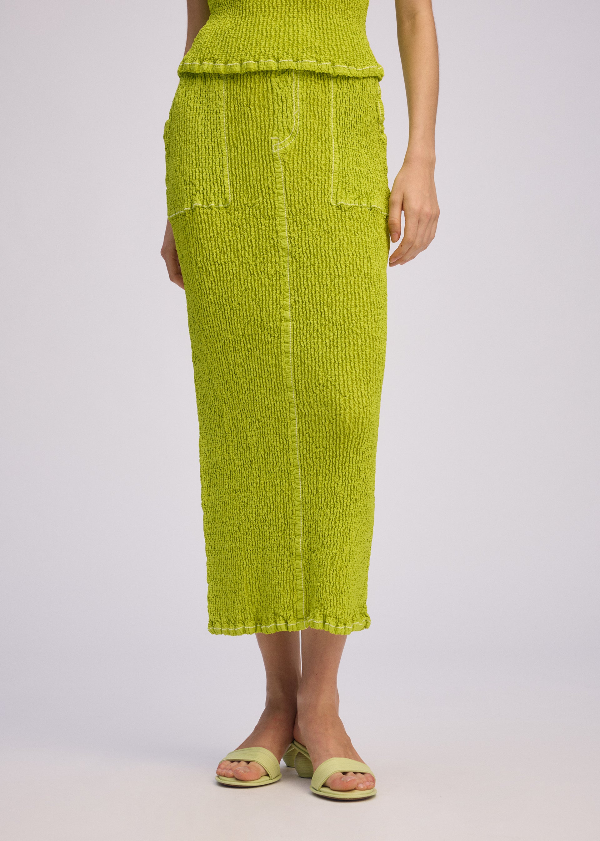 Zoomed view A person wears the ISSEY MIYAKE WAVELET ENSEMBLE—a textured, bright lime green jacquard stretch midi skirt and matching sleeveless top with open-toe sandals; only the lower half is visible against a light background.