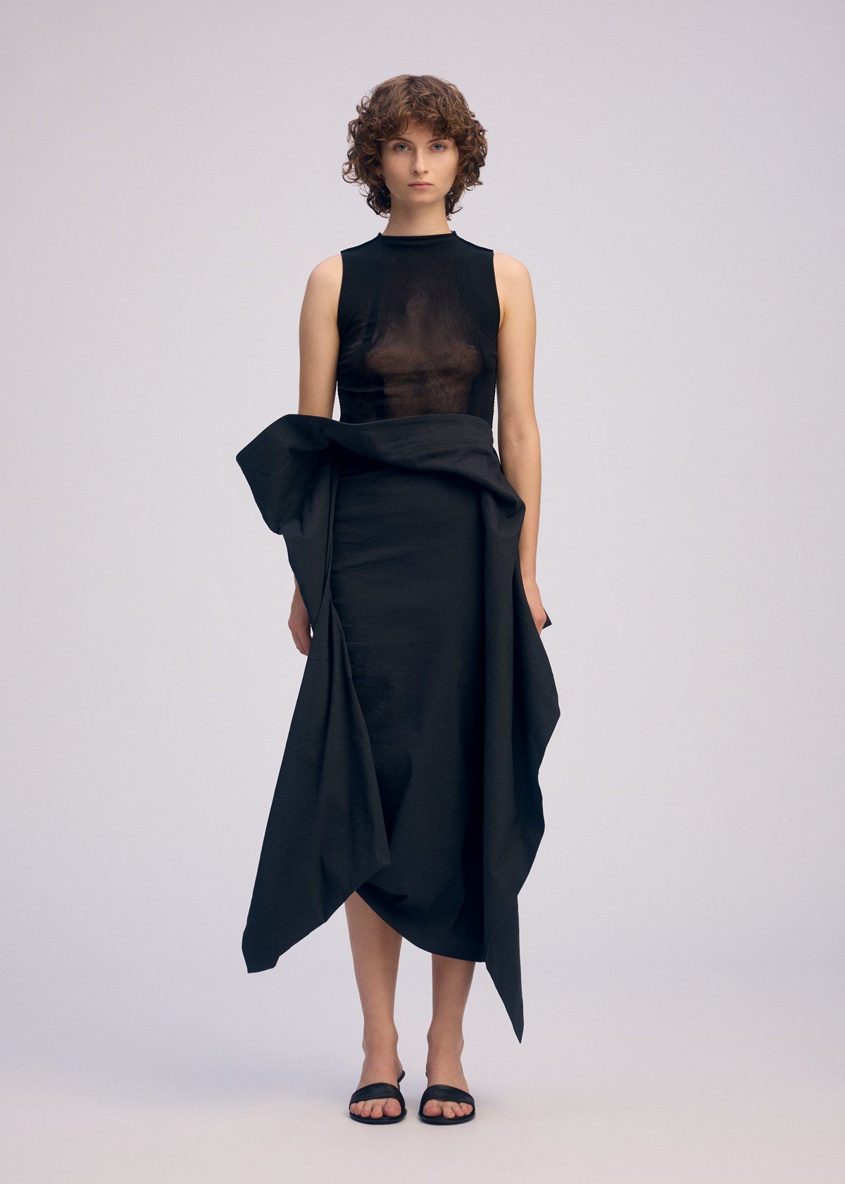 A person with short curly hair wears ISSEY MIYAKE's CONCEALED: a sleeveless black sheer top and an asymmetrical 3D draped skirt, paired with black sandals, standing against a plain light background. view 5 of 7