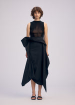 A person with short curly hair wears ISSEY MIYAKE's CONCEALED: a sleeveless black sheer top and an asymmetrical 3D draped skirt, paired with black sandals, standing against a plain light background.