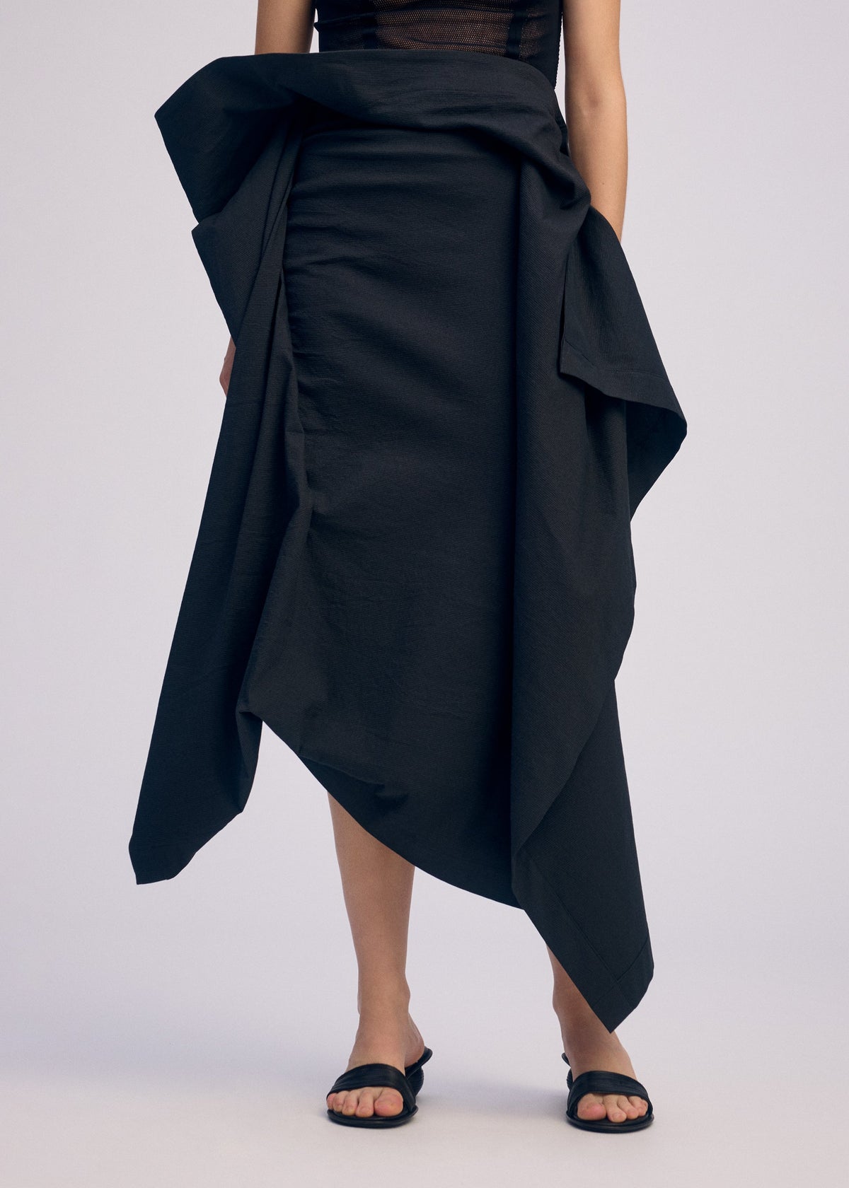 A person stands against a plain light background, wearing ISSEY MIYAKE's CONCEALED—a black, asymmetrical three-dimensional skirt with draped fabric—paired with black sandals. Their upper body is partially visible. view 7 of 7