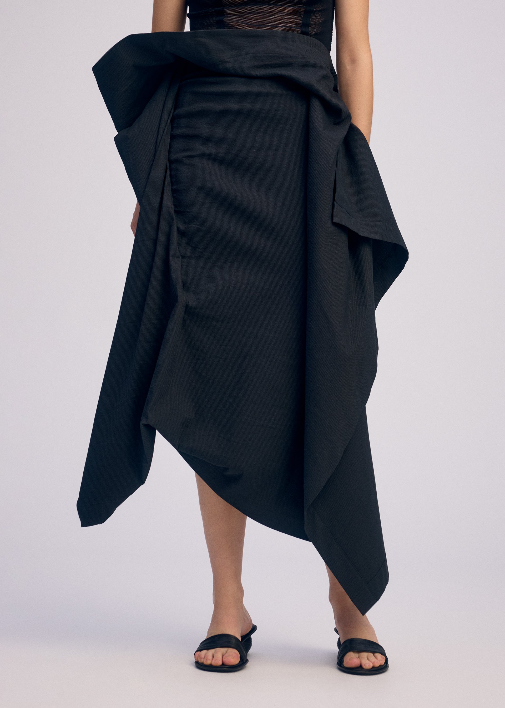 Zoomed view A person stands against a plain light background, wearing ISSEY MIYAKE's CONCEALED—a black, asymmetrical three-dimensional skirt with draped fabric—paired with black sandals. Their upper body is partially visible.