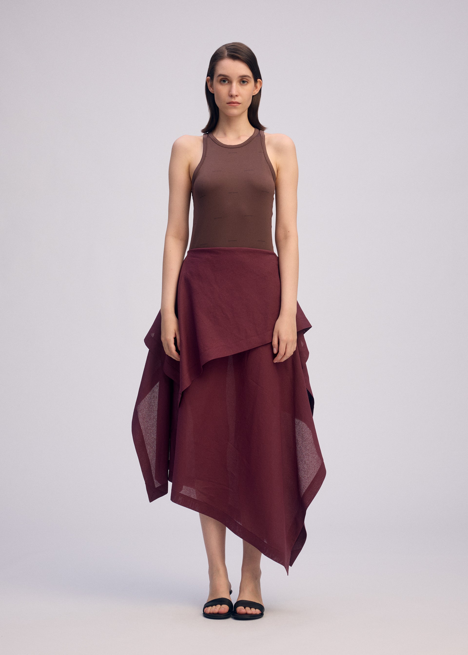 Zoomed view A woman with straight, shoulder-length dark hair stands facing forward, wearing a sleeveless brown top and the ISSEY MIYAKE ALL SQUARES asymmetrical burgundy skirt in ultra-fine polyester. She pairs it with black sandals against a plain light background.