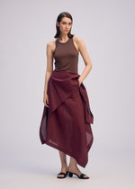 A woman with straight, shoulder-length hair and a neutral expression stands against a plain background wearing ISSEY MIYAKE’s ALL SQUARES brown sleeveless top, a burgundy asymmetric wrap skirt in ultra-fine polyester, and black sandals.