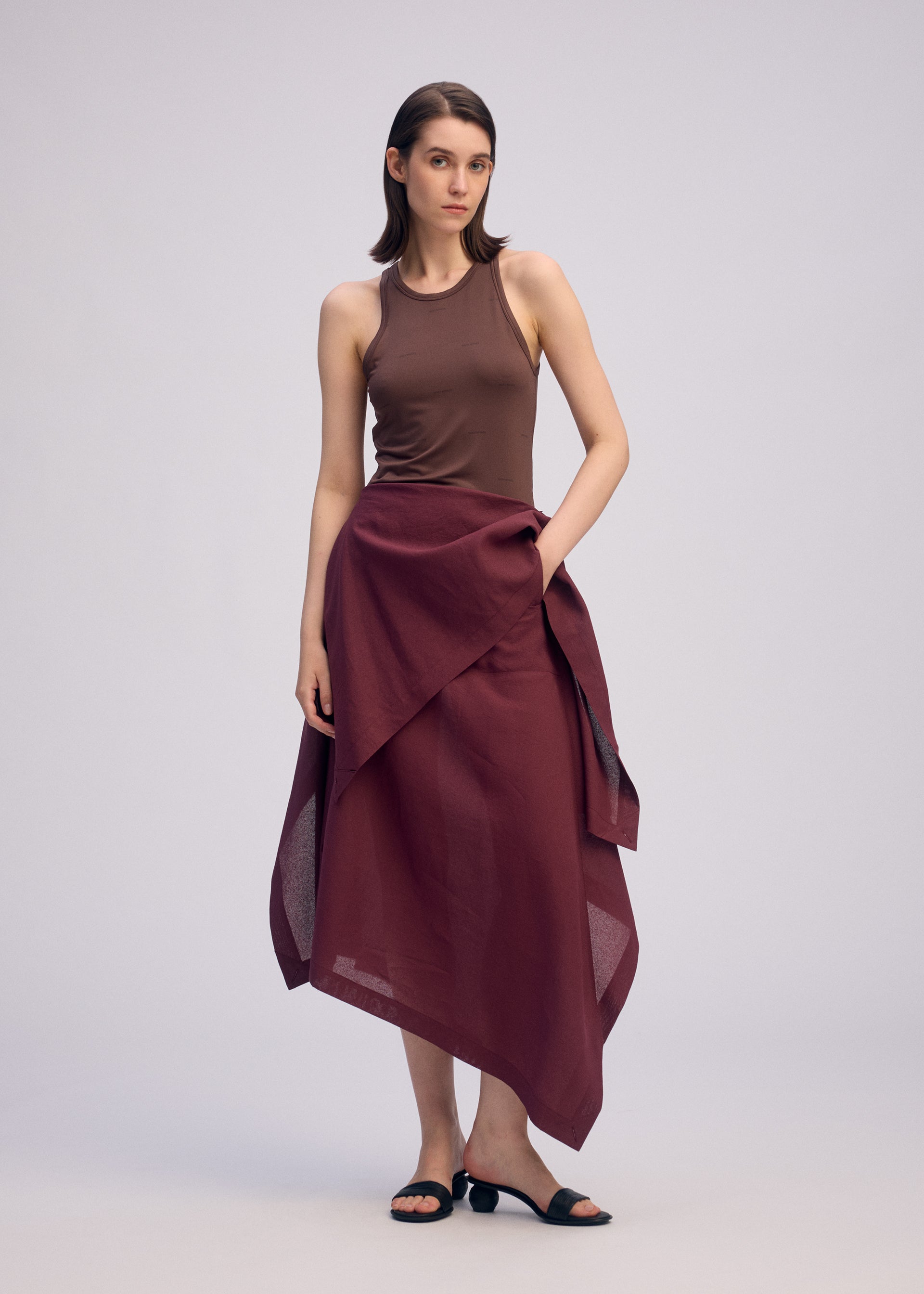 Zoomed view A woman with straight, shoulder-length hair and a neutral expression stands against a plain background wearing ISSEY MIYAKE’s ALL SQUARES brown sleeveless top, a burgundy asymmetric wrap skirt in ultra-fine polyester, and black sandals.