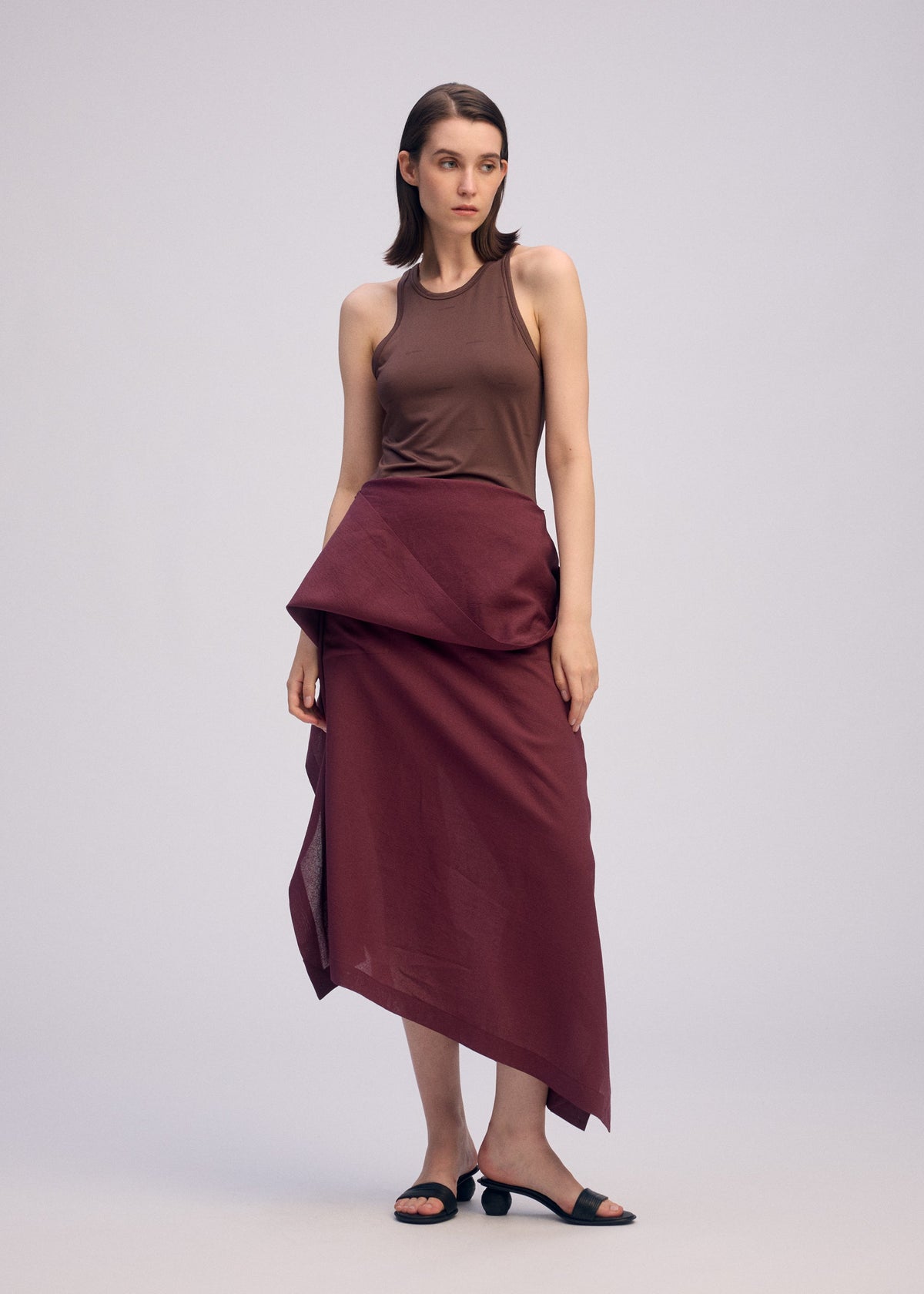 A woman with straight brown hair stands against a plain background wearing ISSEY MIYAKE’s ALL SQUARES sleeveless brown top, a maroon asymmetrical wrap skirt inspired by Japanese Washi paper, and black sandals. She gazes to the side with a neutral expression. view 8 of 10