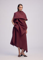 A person stands against a plain background, wearing the ISSEY MIYAKE ALL SQUARES sleeveless, asymmetrical deep burgundy dress with multiple draped layers made from ultra-fine polyester, paired with black heeled sandals.