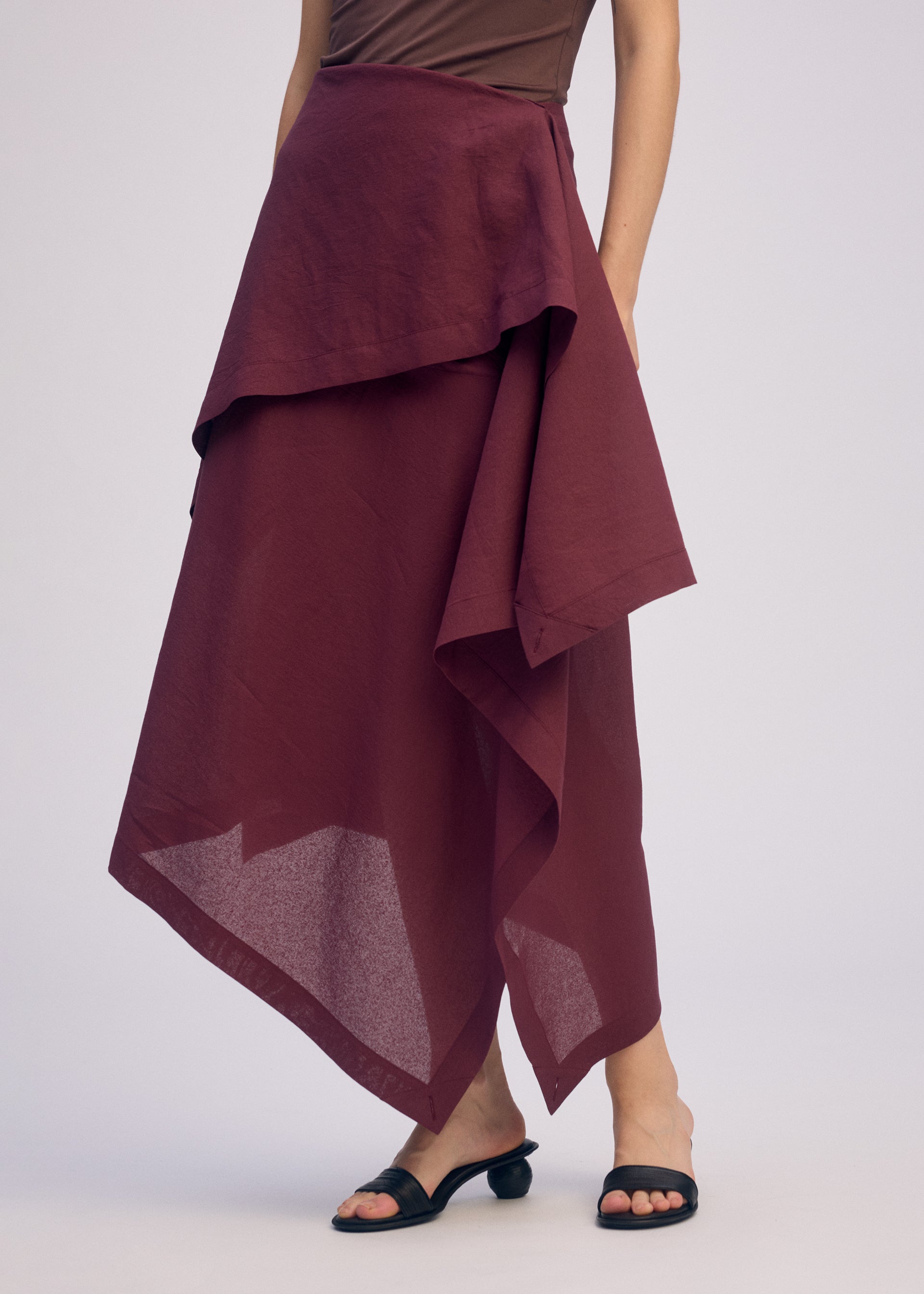 Zoomed view A person models ISSEY MIYAKE's ALL SQUARES in a brown sleeveless top and burgundy asymmetric layered polyester skirt with black open-toe sandals, face hidden to reflect the collection’s minimalist aesthetic against a light backdrop.