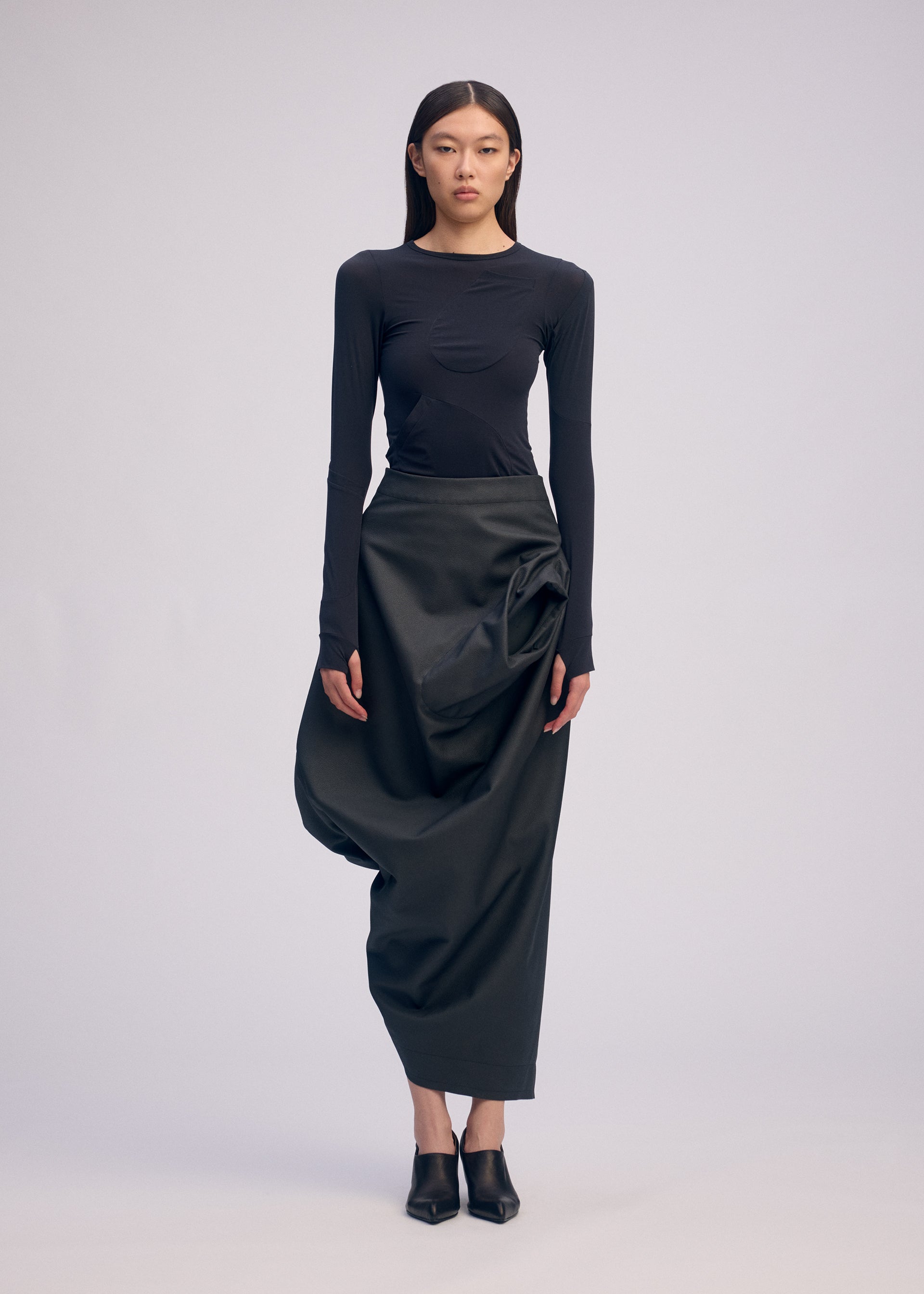 Zoomed view A woman stands against a plain background wearing the ISSEY MIYAKE PEU FORM³ fitted black long-sleeve top and artificial leather skirt from the Autumn Winter 2025/26 Collection, paired with pointed black shoes and her straight dark hair.