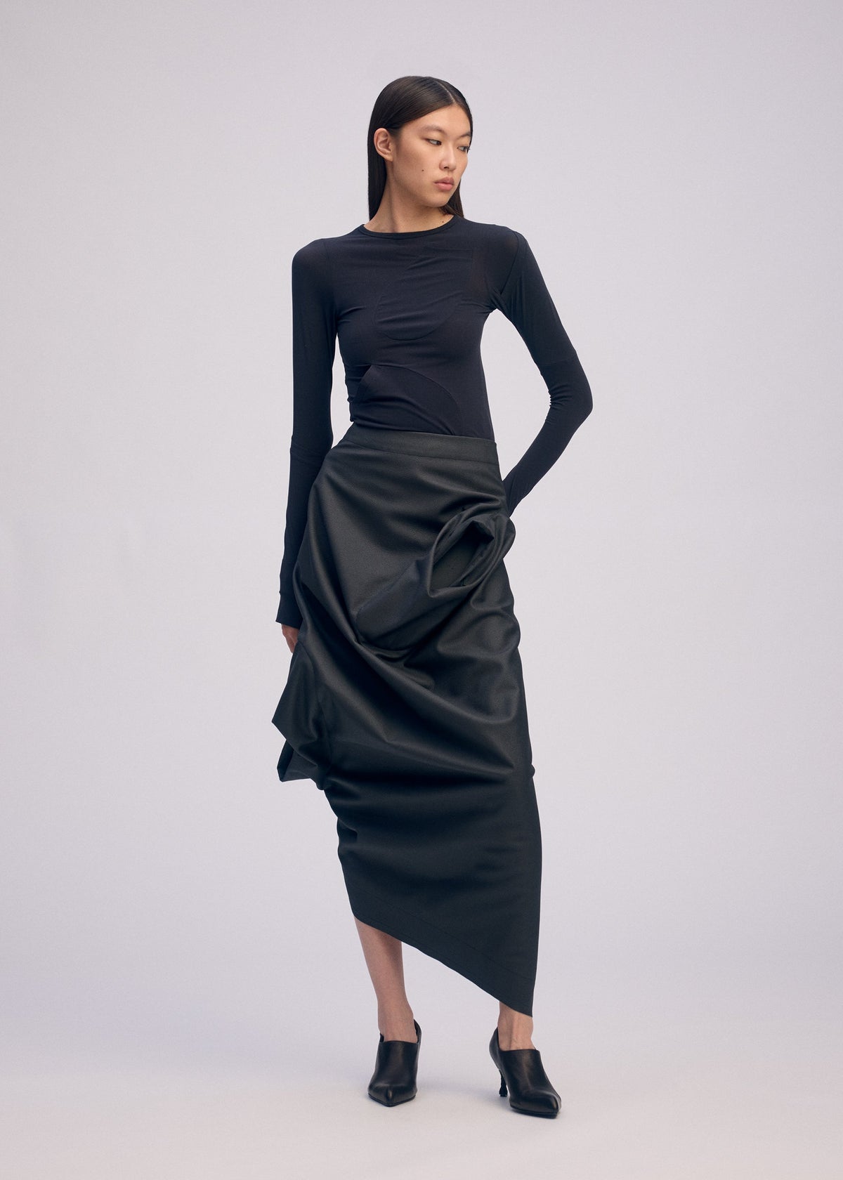 A woman poses against a plain background in a fitted black long-sleeve top, the ISSEY MIYAKE PEU FORM³ asymmetrical artificial leather skirt, and black heeled mules—an elegant look from the Autumn Winter 2025/26 Collection. view 6 of 10