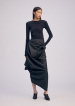 A woman poses against a plain background in a fitted black long-sleeve top, the ISSEY MIYAKE PEU FORM³ asymmetrical artificial leather skirt, and black heeled mules—an elegant look from the Autumn Winter 2025/26 Collection.