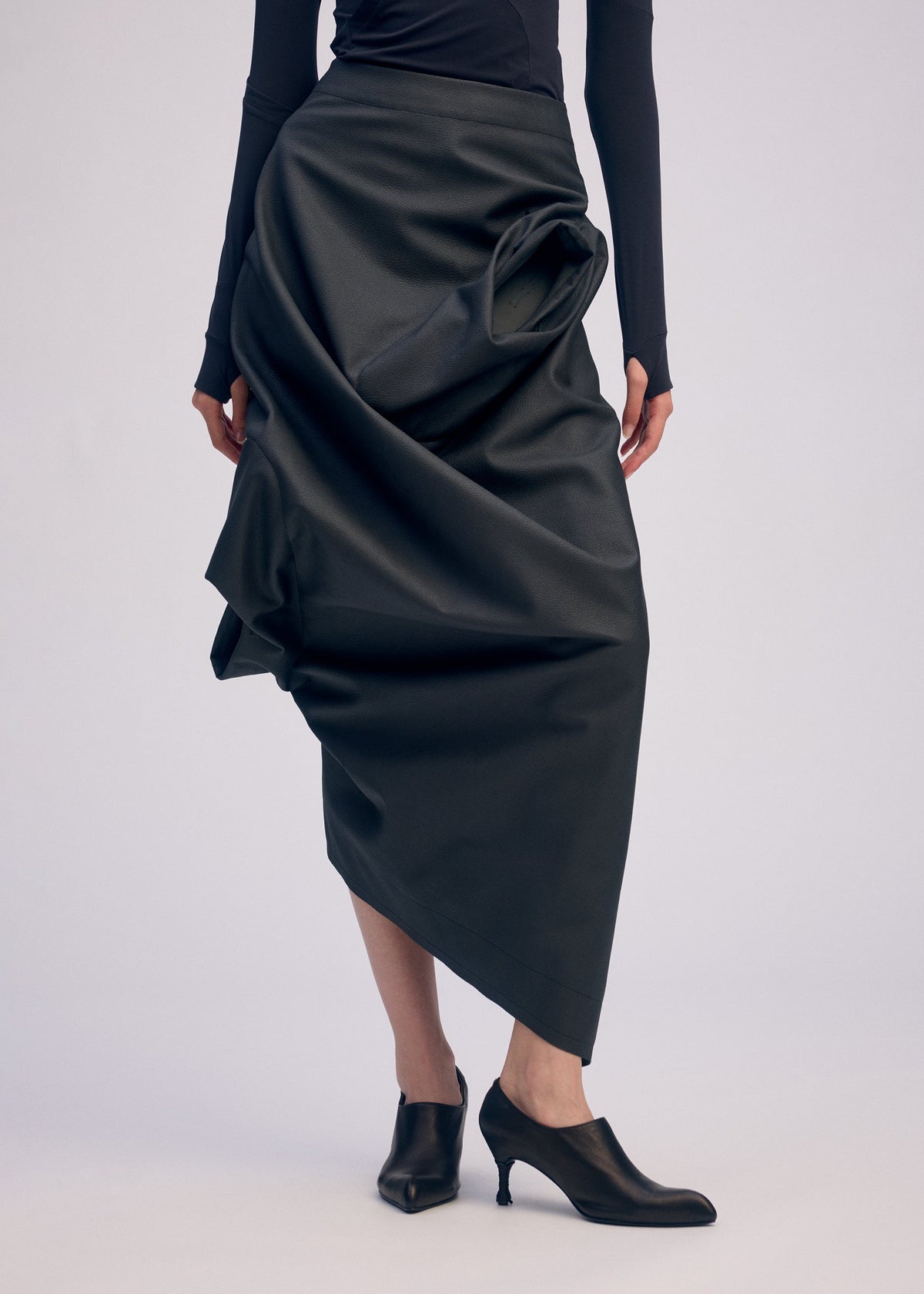 A person wears a long black asymmetrical artificial leather skirt with draped details and a fitted black top from the ISSEY MIYAKE PEU FORM³ Autumn Winter 2025/26 Collection, paired with black heeled shoes against a plain background. view 7 of 10