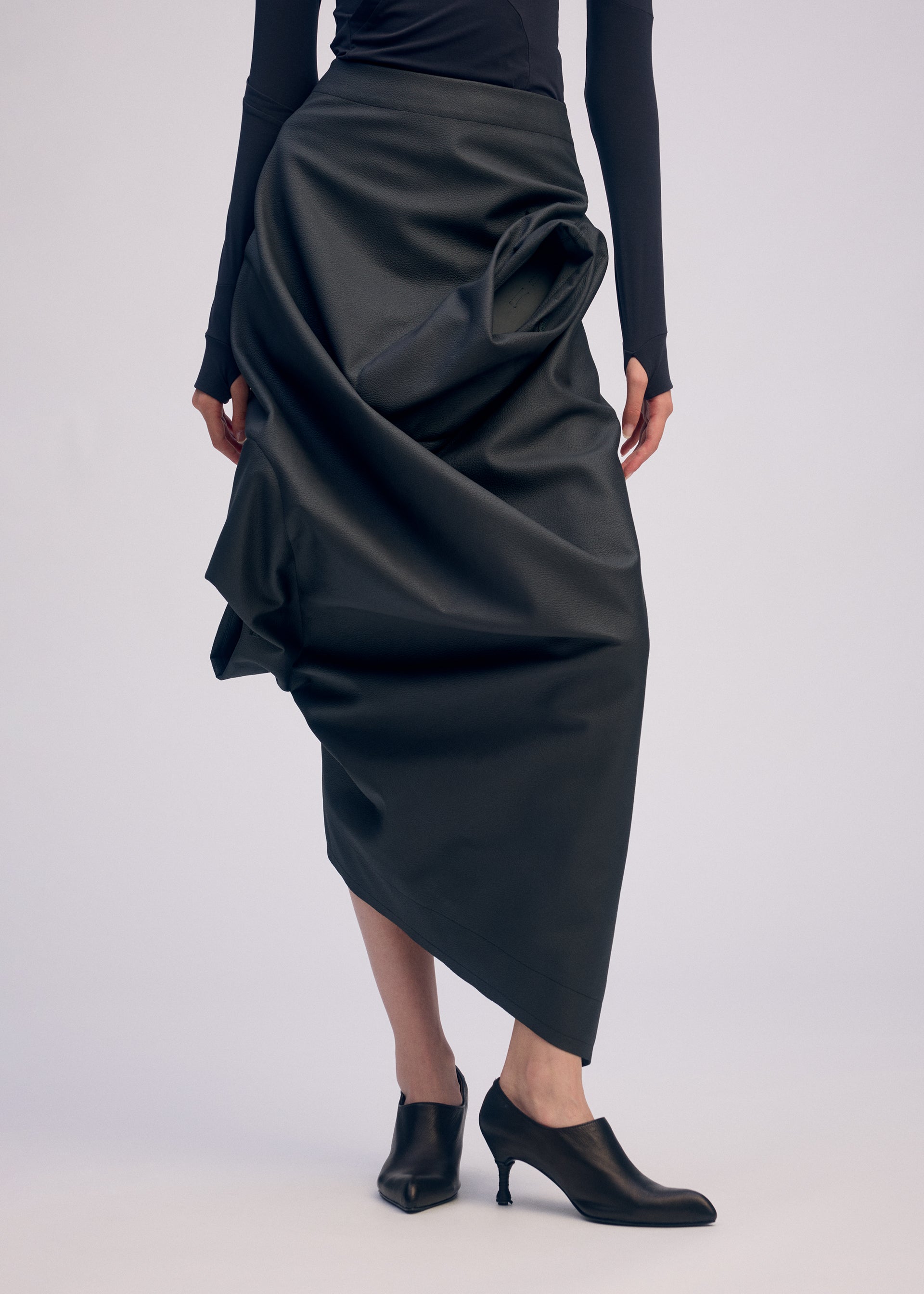 Zoomed view A person wears a long black asymmetrical artificial leather skirt with draped details and a fitted black top from the ISSEY MIYAKE PEU FORM³ Autumn Winter 2025/26 Collection, paired with black heeled shoes against a plain background.