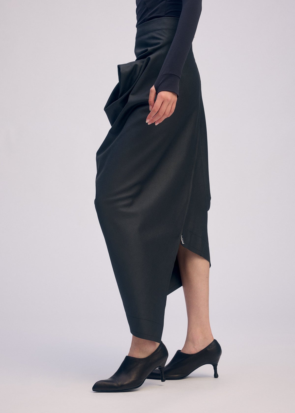 A person wears a long, asymmetrical artificial leather skirt with gathered details, a fitted long-sleeve black top, and black low-heeled shoes from the ISSEY MIYAKE PEU FORM³ Collection, standing against a plain light background. view 8 of 10