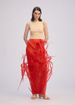 A woman with a serious expression models the ISSEY MIYAKE URBAN JUNGLE: a sleeveless beige top and bright red skirt featuring bold, spiky embellishments, paired with flat sandals against a plain background.