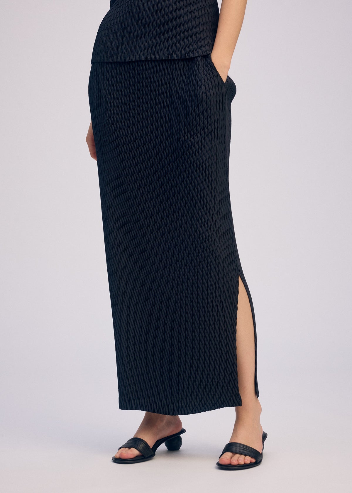 The lower body is shown against a plain background, wearing the ISSEY MIYAKE SLEEK PLEATS long black skirt with side slit, paired with black slide sandals. view 8 of 8