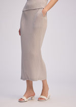 A person wears the ISSEY MIYAKE BOX SHOULDER PLEATS light gray double-pleated midi skirt and matching top, with a hand in their pocket and white open-toe heeled sandals, against a plain light background.