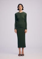 A woman with short dark hair wears the ISSEY MIYAKE RHOMBUS PLEATS—a textured green top and matching ankle-length skirt with a sleek silhouette. She pairs the set with black sandals, standing against a plain light background.