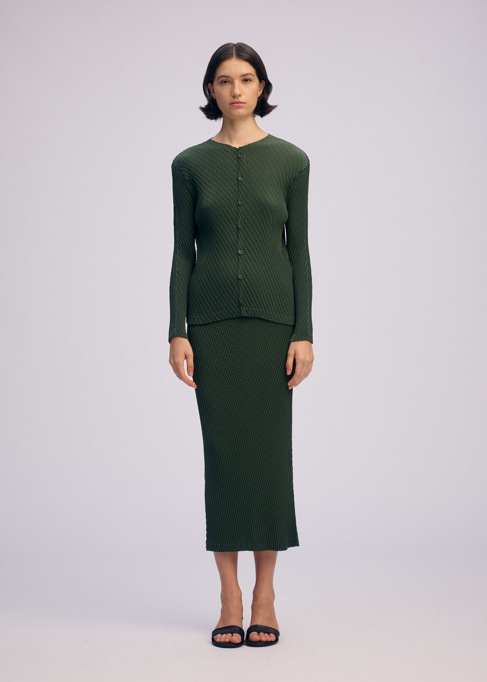 Zoomed view A woman with short dark hair wears the ISSEY MIYAKE RHOMBUS PLEATS—a textured green top and matching ankle-length skirt with a sleek silhouette. She pairs the set with black sandals, standing against a plain light background.