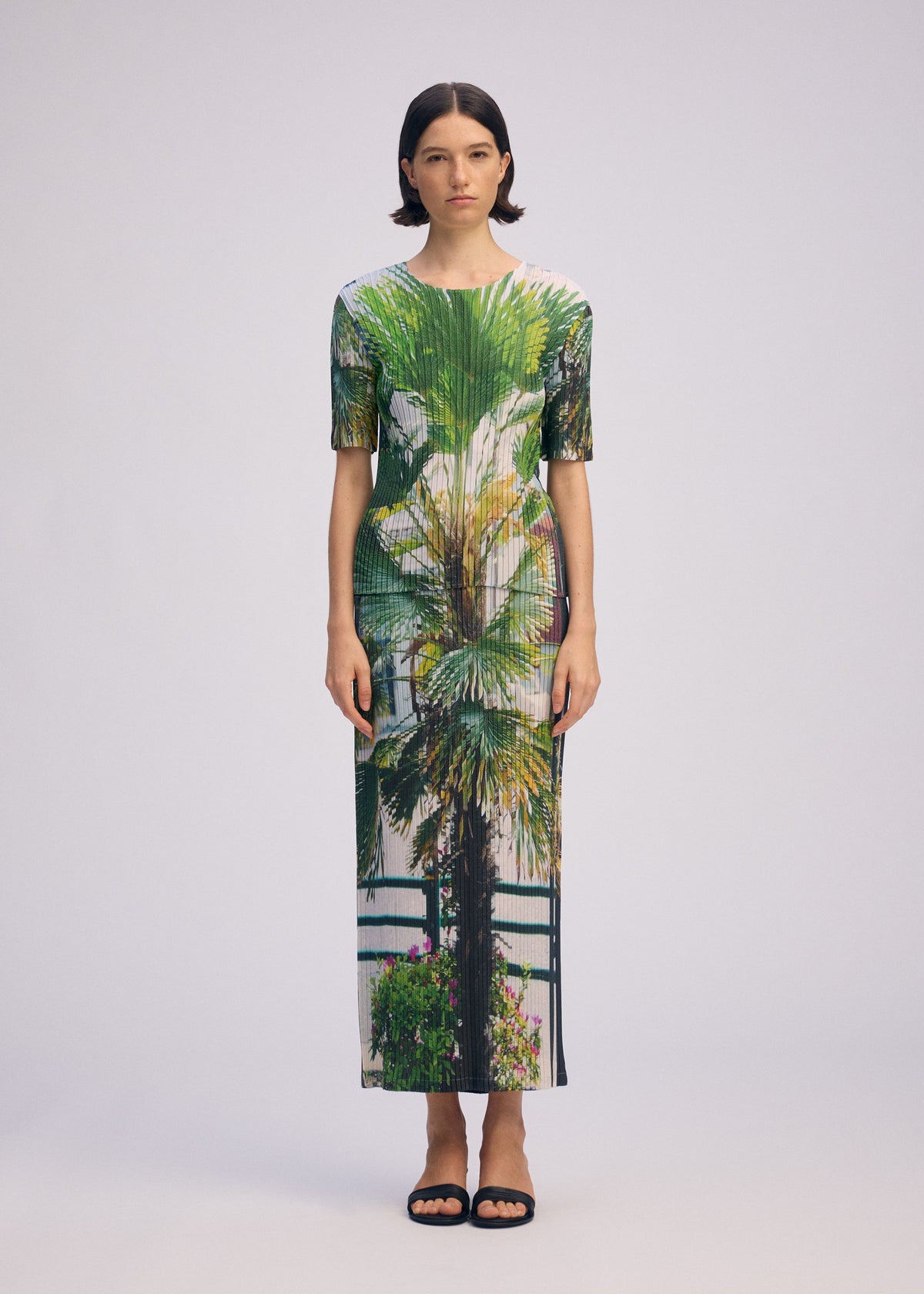 A woman with short dark hair faces forward in a fitted, ankle-length URBAN JUNGLE PLEATS dress by ISSEY MIYAKE, featuring a vibrant palm print. She wears black sandals and stands against a plain light background with a neutral expression. view 5 of 7