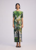 A woman with short dark hair faces forward in a fitted, ankle-length URBAN JUNGLE PLEATS dress by ISSEY MIYAKE, featuring a vibrant palm print. She wears black sandals and stands against a plain light background with a neutral expression.