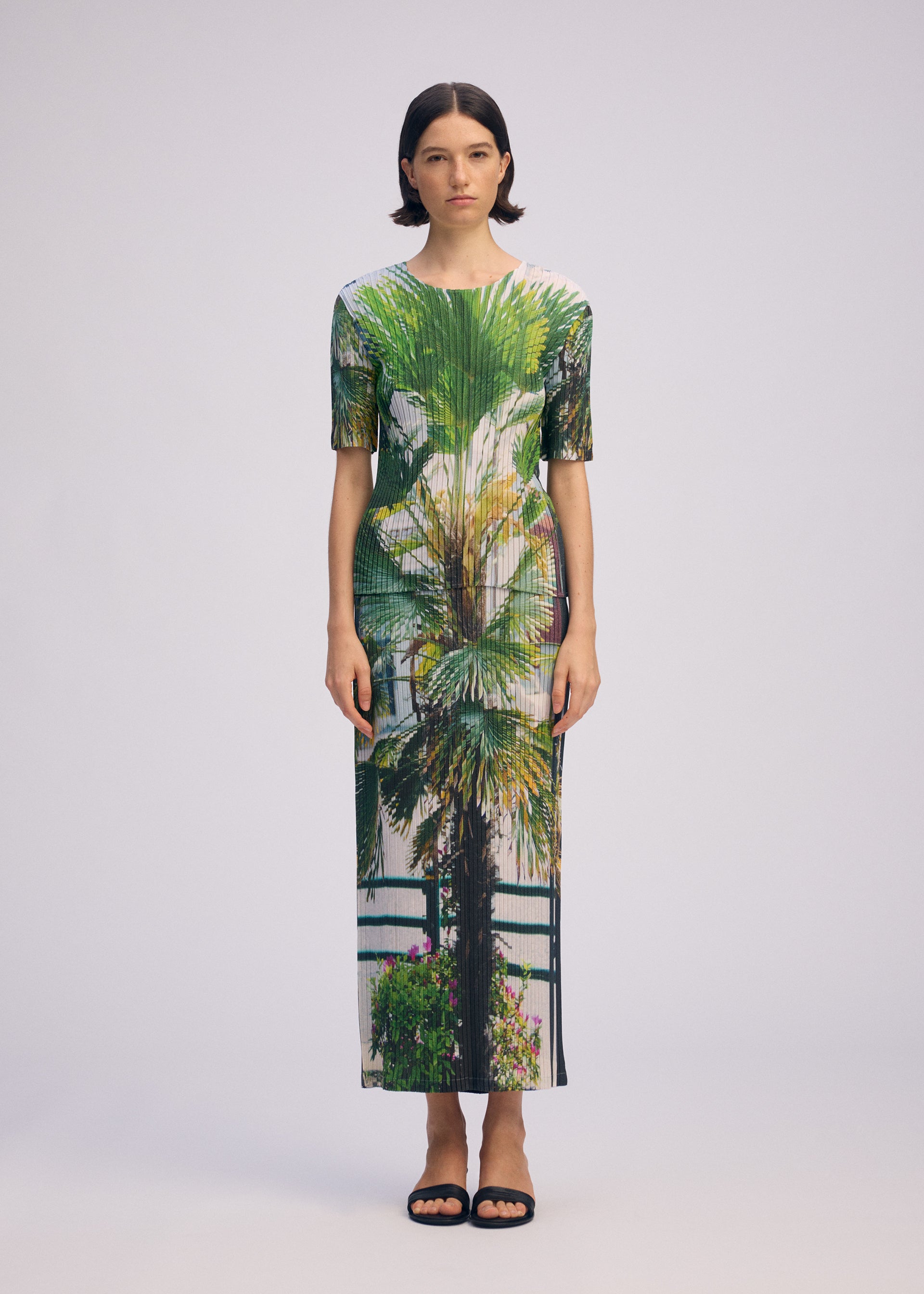 Zoomed view A woman with short dark hair faces forward in a fitted, ankle-length URBAN JUNGLE PLEATS dress by ISSEY MIYAKE, featuring a vibrant palm print. She wears black sandals and stands against a plain light background with a neutral expression.