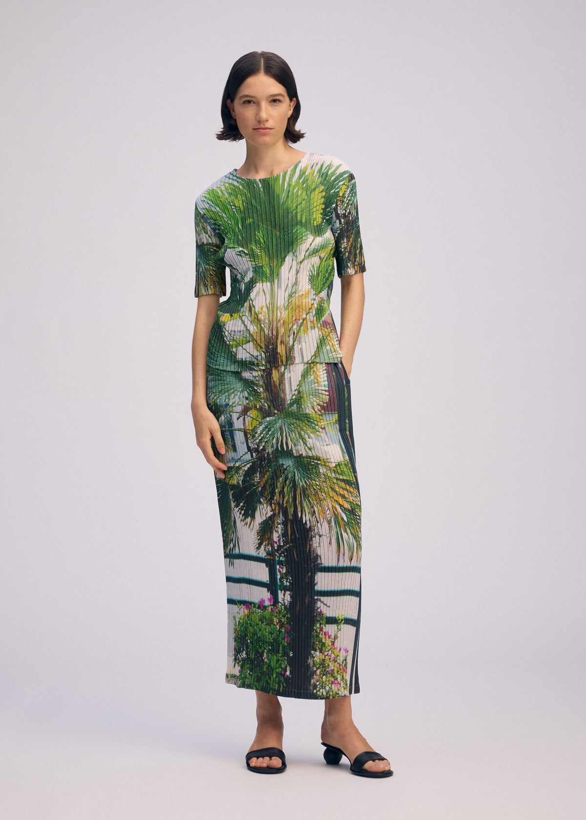 A woman with short dark hair wears the ISSEY MIYAKE URBAN JUNGLE PLEATS dress, featuring a vivid palm tree and greenery print. She pairs it with black sandals in a plain light gray studio background. view 6 of 7
