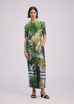 A woman with short dark hair wears the ISSEY MIYAKE URBAN JUNGLE PLEATS dress, featuring a vivid palm tree and greenery print. She pairs it with black sandals in a plain light gray studio background.