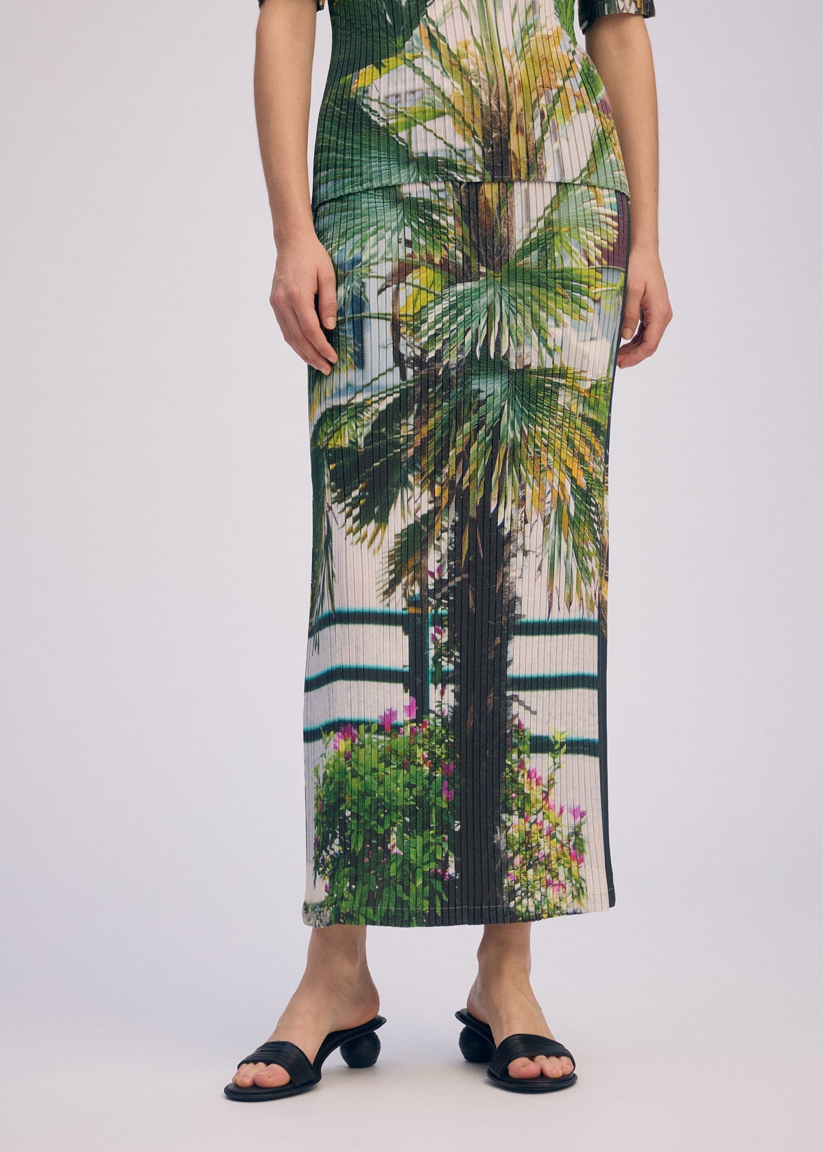 A person wears the ISSEY MIYAKE URBAN JUNGLE PLEATS skirt with a vivid palm and plant print, standing against a plain light background, cropped at the shoulders to show only lower legs and black sandals. view 7 of 7